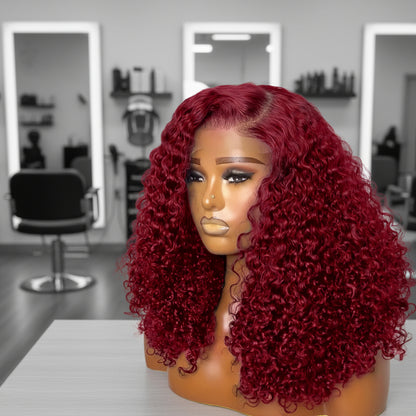 Mannequin head featuring a NuHaire burgundy pixie curly wig, showcasing rich burgundy 99J color, with bouncy, combed-out pixie curls and thin transparent lace options available in 4x4 and 13x4.