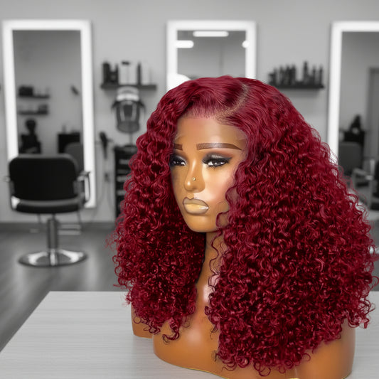 Mannequin head featuring a NuHaire burgundy pixie curly wig, showcasing rich burgundy 99J color, with bouncy, combed-out pixie curls and thin transparent lace options available in 4x4 and 13x4.